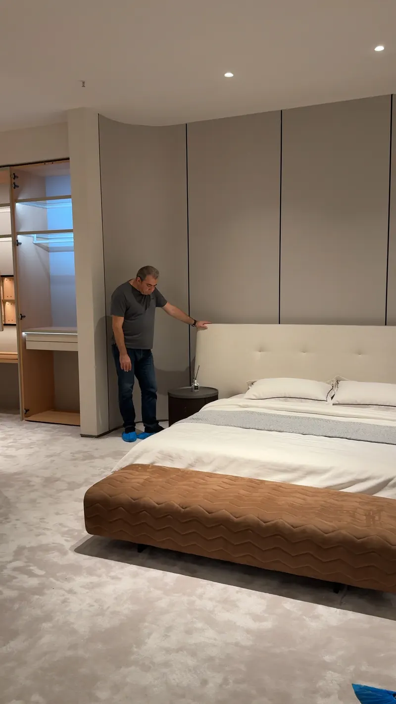 An adult touches a pale upholstered headboard beside a bed in a showroom.