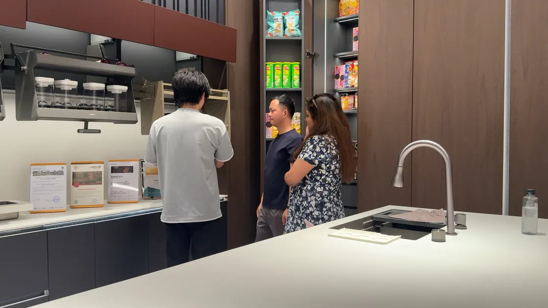Three people stand beside open kitchen storage units and a display counter.
