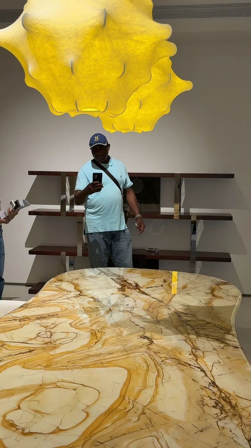 A person stands behind a patterned dining table with a phone, beneath a yellow light fixture.